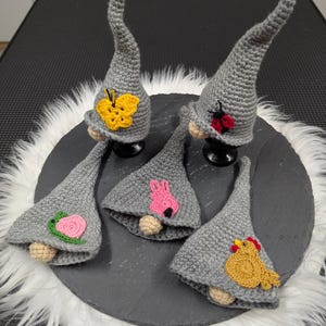 Könnte beinhalten: Eine Sammlung grauer gehäkelter Zwergornamente, jedes mit einem einzigartigen Design. Einer der Zwerge hat einen gelben Schmetterling, ein anderer einen roten Marienkäfer, ein dritter ein rosa Kaninchen und der letzte ein gelbes Huhn. Die Zwerge haben beige Nasen.