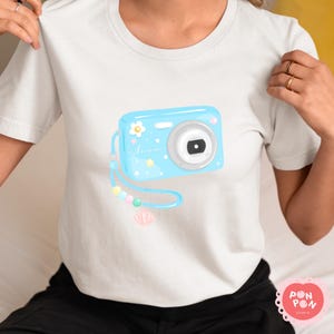 May include: A white short-sleeve t-shirt featuring a graphic of a light blue digital camera. The camera design includes a small white flower, yellow stars, and the word Summer in cursive script. A blue wrist strap with pastel beads and a shell charm.