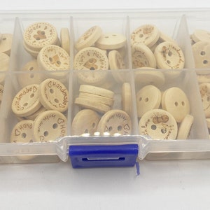 May include: A clear plastic storage box filled with wooden buttons. The buttons are round and have two holes. Some of the buttons have the word "Handmade" written on them.
