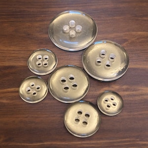 May include: A set of six clear plastic buttons with four holes each. The buttons are arranged in a circle on a wooden surface.