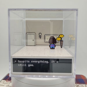 May include: A clear acrylic cube diorama featuring a pixelated character from the video game Undertale. The character is standing in a room with a door, a potted plant, and a wall with a picture. The text "Despite everything, it's still you." is displayed on a black screen at the bottom of the diorama.