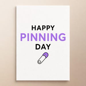 Może przedstawiać: Biała prostokątna kartka z czarnym napisem HAPPY i DAY, z wyśrodkowanym słowem PINNING zapisanym dużymi fioletowymi literami. Poniżej tekstu znajduje się mała ilustracja srebrnej agrafki z fioletową plastikową główką.