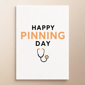 Może przedstawiać: Biała prostokątna kartka z czarnym napisem HAPPY i DAY oraz słowem PINNING zapisanym dużymi pomarańczowymi literami na środku. Poniżej tekstu znajduje się czarno-pomarańczowa graficzna ilustracja stetoskopu medycznego na białym tle.