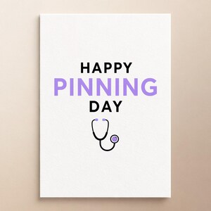 Może przedstawiać: Biała prostokątna kartka z czarnym napisem HAPPY i DAY, z wyśrodkowanym słowem PINNING zapisanym dużymi jasnofioletowymi literami. Pod tekstem znajduje się prosta czarno-jasnofioletowa graficzna ilustracja stetoskopu.