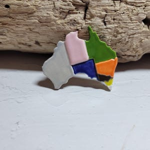 May include: A ceramic pin in the shape of Australia, divided into sections of grey, pink, blue, green, orange, black, and yellow. The pin is set against a light background and a piece of driftwood.