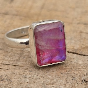 May include: A silver-toned ring featuring a rectangular, faceted gemstone with a vibrant pink and purple iridescent hue. The stone is set in a smooth metal bezel, and the band is a simple, polished silver-colored metal. The text Indiajewellry is visible.