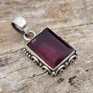 May include: A rectangular, deep red gemstone pendant with a silver-colored metal setting. The pendant has an ornate, detailed border and a small loop at the top for a chain. The gemstone is a rich, dark red color.