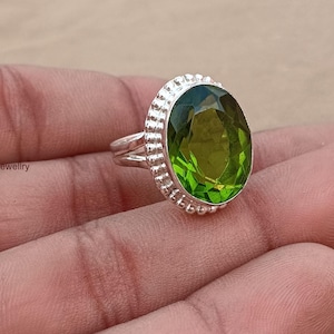 Puede incluir: Un anillo de plata con una gran piedra preciosa de color verde oliva translúcido y corte ovalado. El engaste tiene un borde decorativo de cuentas alrededor de la piedra y una banda dividida. El texto "indiajewellry" es visible a la izquierda.