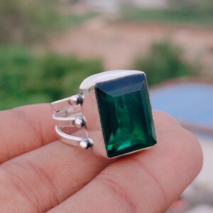 May include: A silver ring featuring a large, rectangular, emerald-green gemstone. The ring has a simple band with three smaller decorative elements on each side of the gemstone. The setting is a bright, polished silver.
