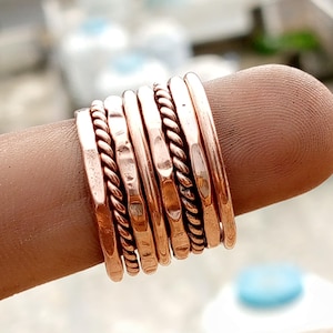 Puede incluir: Una colección de anillos de color cobre, algunos con texturas martilladas y otros con diseños de cuerda trenzada. Los anillos están apilados en un dedo, mostrando sus diversos estilos y acabados. Los anillos tienen un tono metálico cálido.