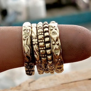 May include: A collection of six gold-colored rings, each with a unique embossed design. The rings are stacked together, showcasing floral, geometric, and textured patterns. The rings are displayed on a finger.