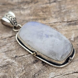 May include: A rectangular moonstone pendant set in a silver bezel with a decorative bail. The gemstone displays a milky, translucent white appearance with subtle blue iridescence. The metal setting features a polished finish and a textured bail.