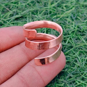 Peut inclure: Une bague en métal de couleur cuivre présentant un design en spirale avec deux extrémités ouvertes. L'anneau possède une finition lisse et polie ainsi qu'une structure plate en forme de ruban qui s'enroule pour créer un aspect double boucle.
