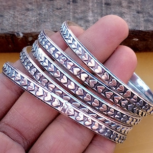 May include: A set of four silver-toned metal bangles, each featuring a continuous embossed pattern of small hearts. The bracelets have a polished finish with darkened recessed areas that highlight the heart shapes along the entire outer surface.