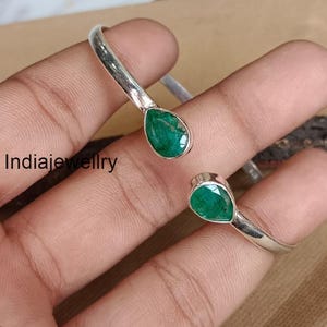 May include: A silver bracelet featuring two teardrop-shaped, vivid green gemstones. The open design allows for size adjustment. The gemstones are set in a bezel setting. The bracelet is presented against a neutral backdrop. The bracelet is a piece of jewelry.