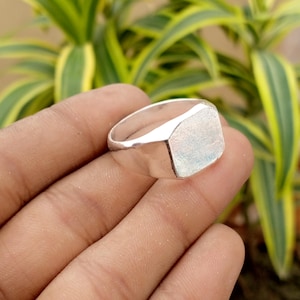 Puede incluir: Un anillo de sello de plata pulida que presenta una cara rectangular plana con esquinas redondeadas y una banda lisa y reflectante. La superficie metálica exhibe un brillo metálico, destacando las líneas limpias y la construcción simple de la pieza.