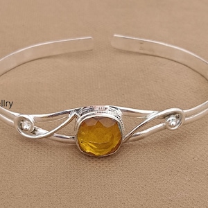 May include: A silver cuff bracelet featuring a central faceted amber-coloured gemstone. The metal band has a polished finish with decorative swirling wire details on each side of the stone. The text IndiaJewellry is visible on the left side.
