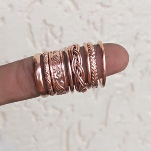 May include: A set of seven copper rings featuring various designs including a smooth band, a floral embossed band, a braided band, and a leaf-patterned band. The rings are arranged in a row, showcasing different textures and widths.