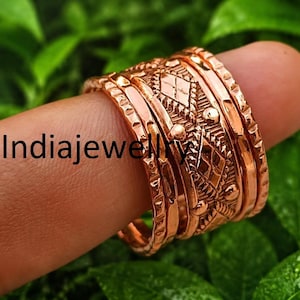 Op de afbeelding: Een brede koperen ring met een centrale band voorzien van een reliëfpatroon van ruiten en stippen, geflankeerd door twee dunnere banden met getextureerde randen. Het metaal heeft een warme koperkleur. De tekst Indiajewellery is zichtbaar.