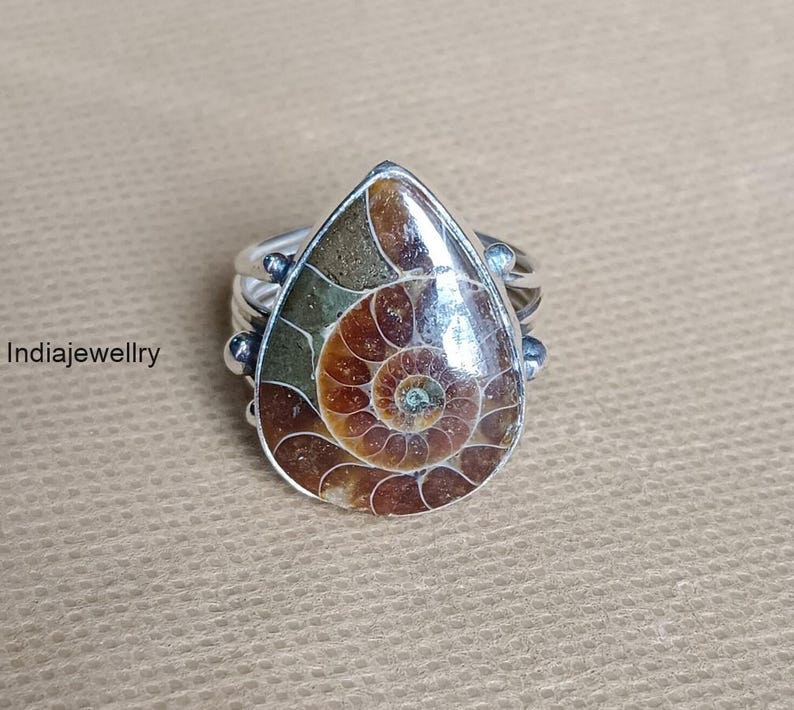 May include: A silver ring featuring a teardrop-shaped fossilized ammonite. The fossil displays a spiral pattern in shades of brown, tan, and green, set in a silver band. The ring is a unique piece of jewelry.