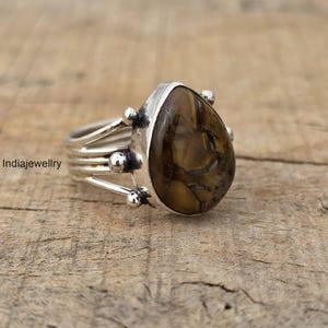 Pode incluir: Anel de prata com pedra olho de tigre em forma de gota. A pedra apresenta tons de castanho e dourado. O anel tem um design multibanda com pequenos detalhes de bolas prateadas.