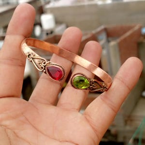 Puede incluir: Brazalete abierto de color cobre con detalles ornamentados. El brazalete presenta una piedra preciosa roja en forma de lágrima en un extremo y una piedra preciosa verde en el otro. Las piedras están engastadas en biseles decorativos.