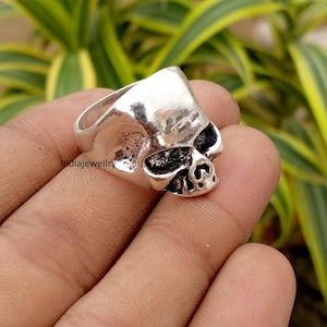 May include: A silver-toned metal ring shaped like a human skull. The ring features a polished, smooth upper cranium surface and a darkened, recessed area around the eye sockets and nasal cavity, creating a high-contrast, detailed skull design.