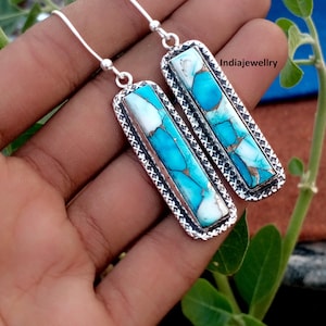 Puede incluir: Un par de pendientes rectangulares colgantes con incrustaciones de piedra turquesa y blanca con vetas de cobre. Las piedras están engastadas en un marco de plata texturizada con un borde decorativo. Los pendientes cuelgan de ganchos de plata e incluyen el texto Indiajewellry.