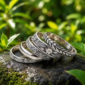 Puede incluir: Un conjunto de cinco anillos de metal plateado apilados. Cada anillo presenta un patrón en relieve distinto, que incluye líneas geométricas, una superficie lisa pulida, motivos florales intrincados y un diseño de hojas repetidas.