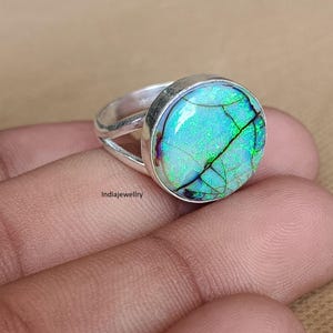 May include: A silver ring featuring a round, iridescent stone with a cracked texture displaying vibrant green and blue flashes. The metal band has a split design, and the text Indiajewellry is visible on the side of the setting.