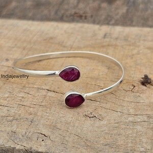 May include: A silver-toned cuff bracelet with two teardrop-shaped, deep red gemstones. The bracelet is open-ended, with the gemstones at each end. The bracelet is displayed on a textured, light brown wooden surface.