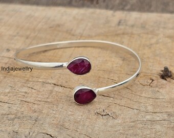 Puede incluir: Un brazalete de plata con dos piedras preciosas rojas oscuras en forma de lágrima. El brazalete abierto tiene una piedra preciosa en cada extremo. El brazalete se presenta sobre una superficie de madera marrón claro texturizada.