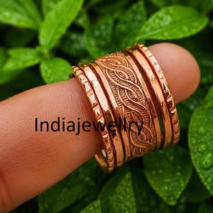 Op de afbeelding: Een set koperkleurige metalen ringen met een centrale band met gevlochten textuur, geflankeerd door twee dunnere banden met een ingekerfd randpatroon. Het woord Indiajewellery staat in zwarte letters in het midden van de ringen.
