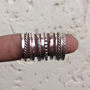 May include: A collection of ten silver rings arranged in a row, featuring various textures and patterns including twisted rope designs, smooth polished bands, beaded details, and intricate engraved geometric motifs on each individual ring.