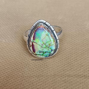 May include: A silver ring featuring a pear-shaped gemstone with a vibrant display of colors, including green, purple, and white. The gemstone is set in a textured silver bezel. The ring is a piece of jewelry.