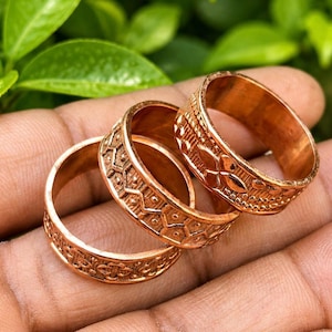 May include: Three copper-colored metal rings featuring distinct embossed patterns. The rings display geometric shapes, including repeating diamond, chevron, and floral-like motifs, with a metallic finish and a consistent band width across all three pieces.