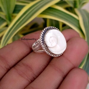 May include: A silver ring featuring a smooth, oval-shaped white shiva eye shell centrepiece. The shell is set within a twisted rope-style metal border. The band is split into two sections near the setting. The text Indiajewellry is visible.