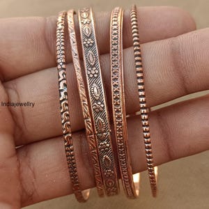 Könnte beinhalten: Ein Set aus fünf Kupferarmreifen mit verschiedenen geprägten Mustern, darunter geometrische Formen, florale Motive und strukturierte Rillen. Das Metall hat ein warmes, rötlich-braunes Finish mit abgedunkelten Vertiefungen zur Betonung.