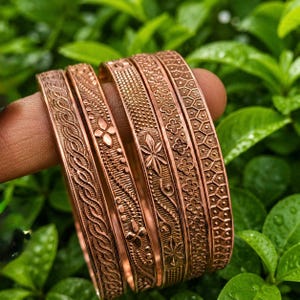 Puede incluir: Un conjunto de cinco brazaletes de metal color cobre, cada uno con un patrón en relieve distinto. Los diseños incluyen cuerda trenzada, pequeños motivos florales, texturas punteadas, formas de hojas estilizadas y un patrón geométrico de panal.