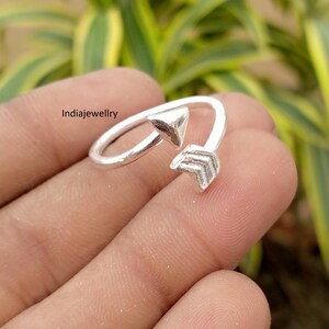 May include: A silver-toned open ring featuring an arrow design with a triangular arrowhead on one end and a chevron-patterned fletching on the other. The text Indiajewellry is visible on the left side of the ring band against a blurred background.