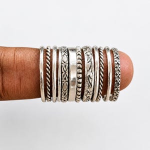 May include: A collection of eleven silver rings featuring various textures and patterns, including smooth bands, twisted rope designs, geometric engravings, and floral motifs, all arranged side-by-side in a row on a finger.