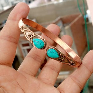May include: Copper cuff bracelet with two turquoise teardrop-shaped stones. The bracelet has ornate silver-colored accents around the stones and a polished copper band. The turquoise stones have a mottled appearance.