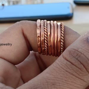 Pode incluir: Uma pilha de nove anéis de cobre, cada um com uma textura diferente, incluindo martelado, torcido e liso. Os anéis são usados em um dedo.