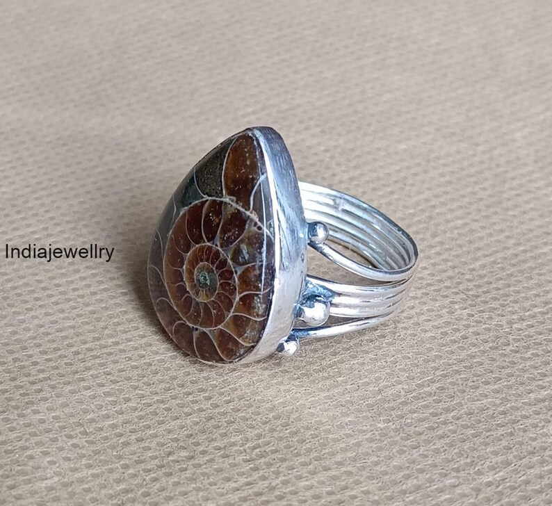 May include: A silver ring featuring a teardrop-shaped, brown and tan fossilized ammonite stone. The ring has a multi-banded silver band with small decorative silver beads. The text "Indiajewellry" is visible on the left side.