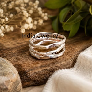 Puede incluir: Un anillo de plata con un diseño de múltiples bandas con una textura irregular, ondulada y martillada. La superficie metálica tiene un acabado pulido y reflectante. El texto Indiajewellry es visible en letras negras sobre el anillo.