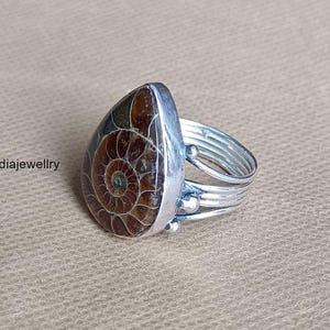 May include: A silver ring with a teardrop-shaped, brown and tan fossilized ammonite stone. The ring band has multiple silver bands and small silver beads. The text "Indiajewellry" is visible on the left side of the image.