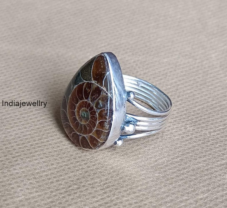 May include: A silver ring with a teardrop-shaped, brown and tan fossilized ammonite stone. The ring band has multiple silver bands and small silver beads. The text "Indiajewellry" is visible on the left side of the image.