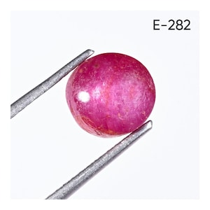 Könnte beinhalten: Ein runder, polierter Rubin-Edelstein, der von einer Metallpinzette vor einem weißen Hintergrund gehalten wird. Der Rubin hat eine tiefrosa-rote Farbe mit einer glatten, leicht strukturierten Oberfläche. Der Text "E-282" befindet sich in der oberen rechten Ecke.