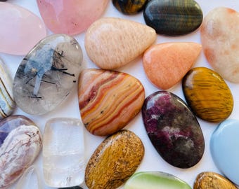 Peut inclure: Une collection de pierres précieuses polies de différentes formes et couleurs. L'assortiment comprend du quartz rose, de l'œil de tigre et des pierres avec des motifs rayés, mouchetés et translucides. Les pierres sont présentées sur une surface blanche.