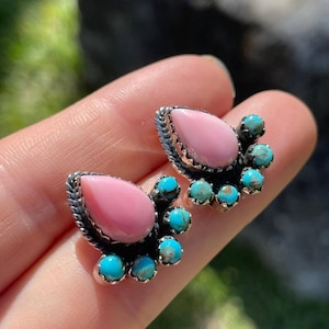 May include: A pair of stud earrings featuring a central teardrop-shaped pink stone surrounded by a silver setting with a twisted rope border. Each earring is accented with five small, round, blue-green turquoise stones along the outer edge.